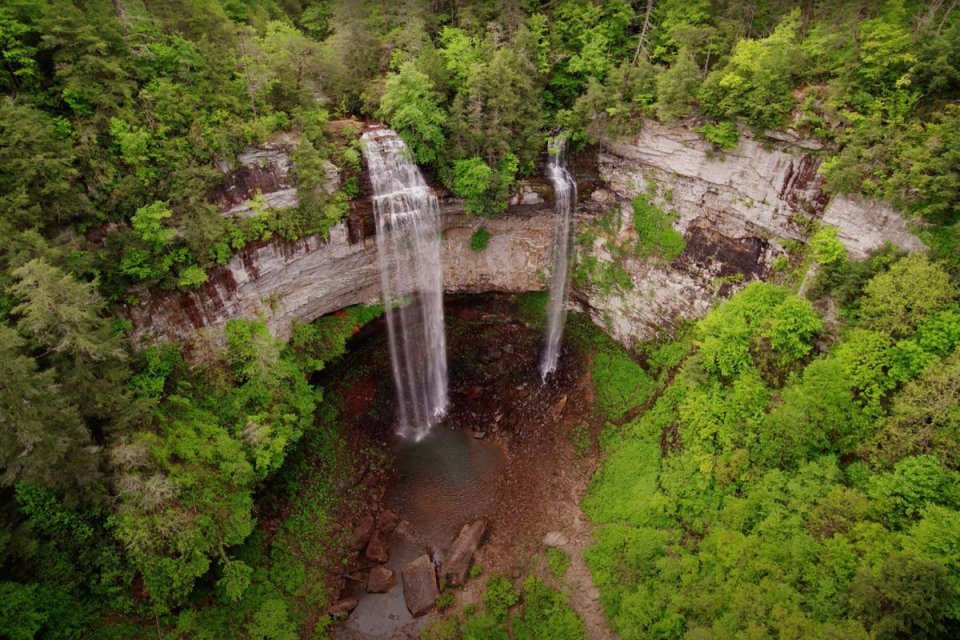 Aerial view of a double waterfall flowing over an ancient bridge in a lush forest.