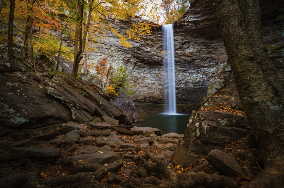 Autumn waterfall cascading into a serene pond surrounded by rocky cliffs and colorful leaves.