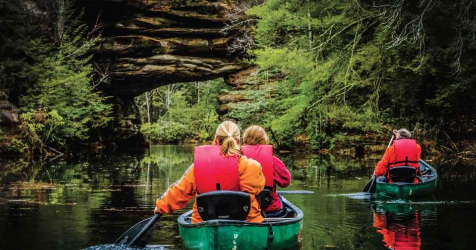 People in bright life jackets kayaking under a natural rock arch surrounded by lush greenery.
