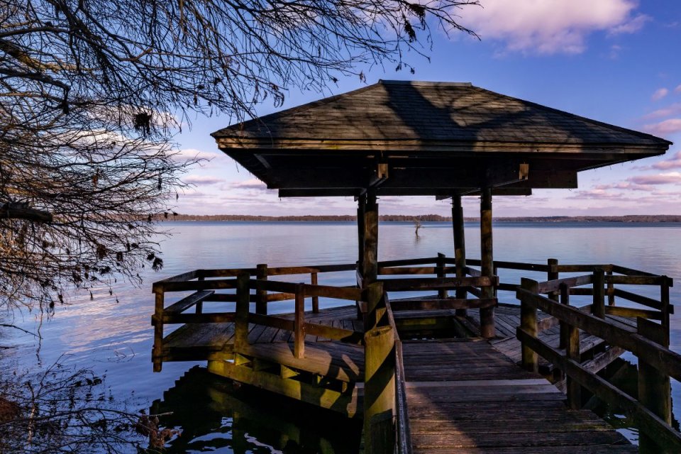 Wooden gazebo on a pier overlooking a tranquil lake at sunset.