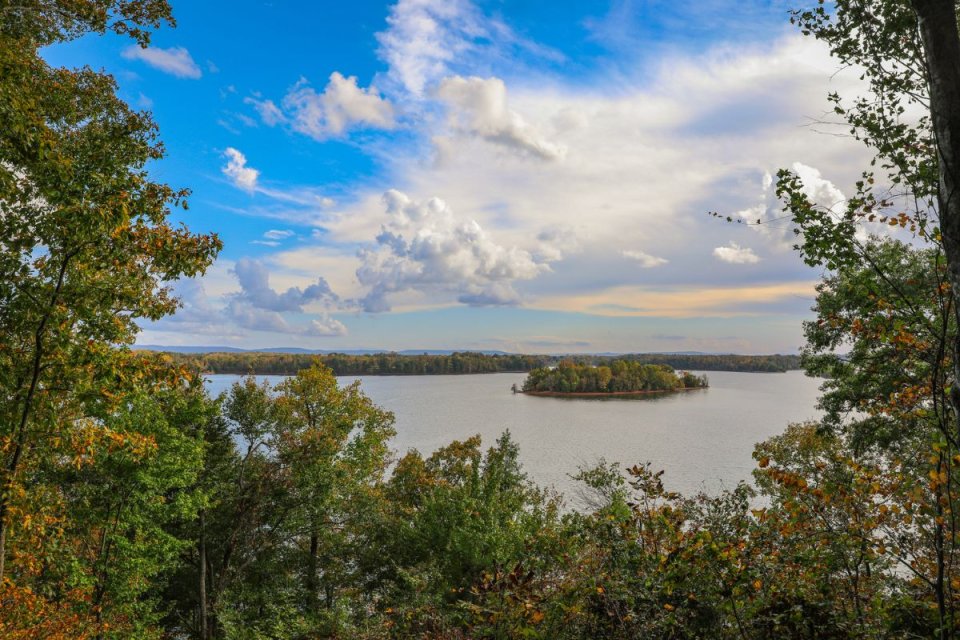 Scenic view of a lake with a small island, surrounded by autumn trees under a cloudy sky.