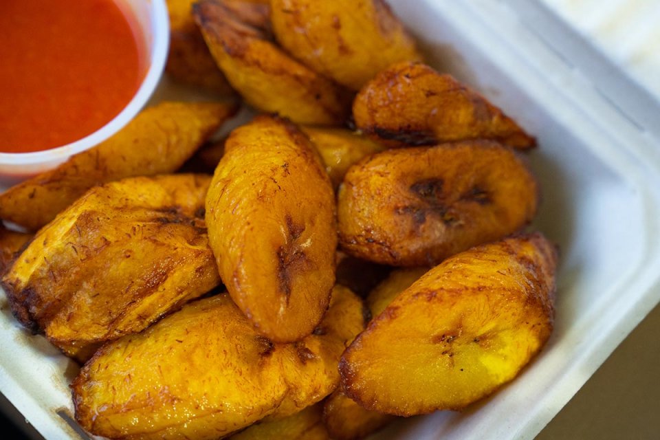 Fried plantains in a takeout container with a side of red sauce.