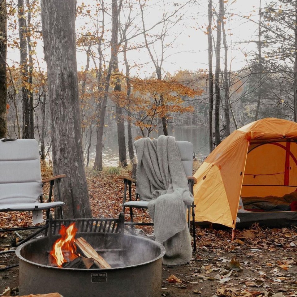 two cushioned camping chairs next to a orange tent and a fire pit in the woods