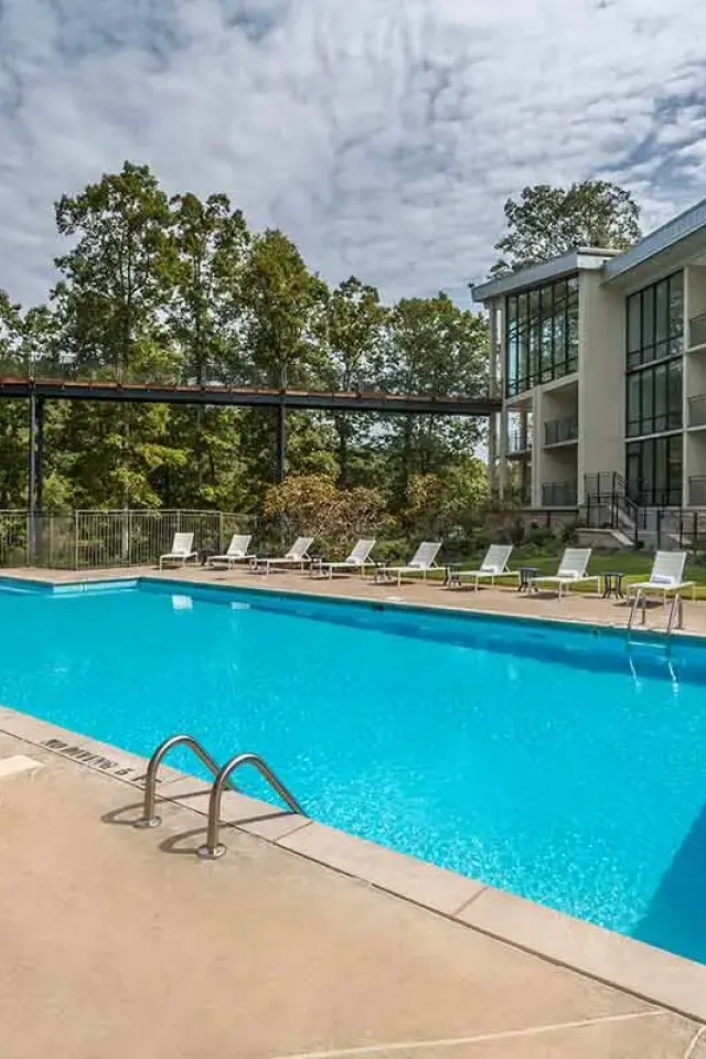 Outdoor pool with loungers, near modern building and trees under a cloudy sky.