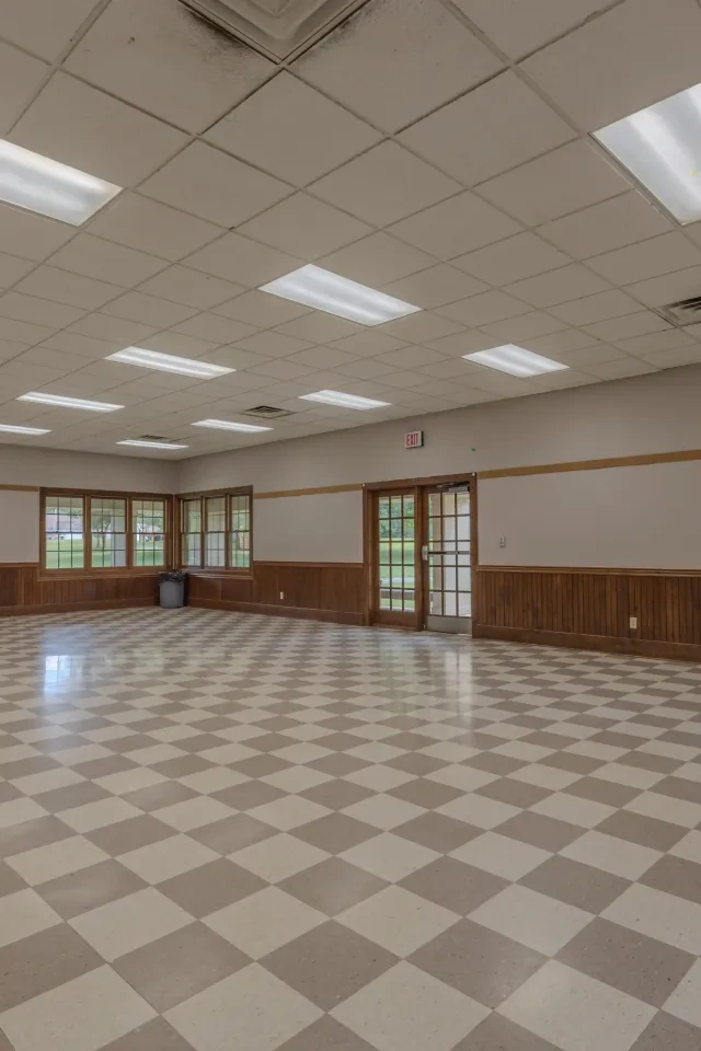 Large empty room with checkered floor and wooden panels, windows, and exit signs.