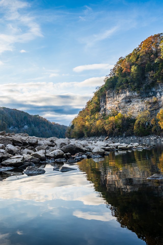 A tranquil river reflects the blue sky and surrounding cliffs, with scattered rocks lining the shore.