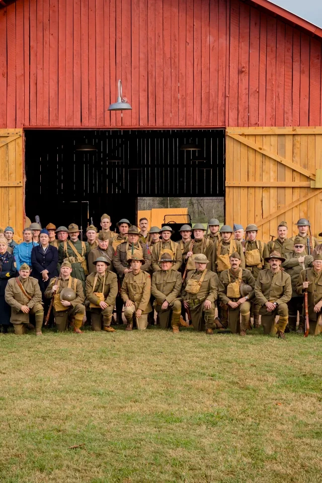 Group photo at a vintage reenactment with people in historical military uniforms and dresses in front of a large red barn, conveying nostalgia.