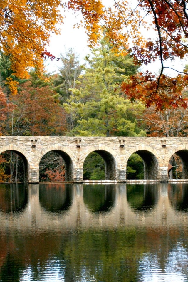 Arched stone bridge spanning a calm lake, surrounded by vibrant fall foliage in shades of orange, yellow, and green, with reflections in the water.