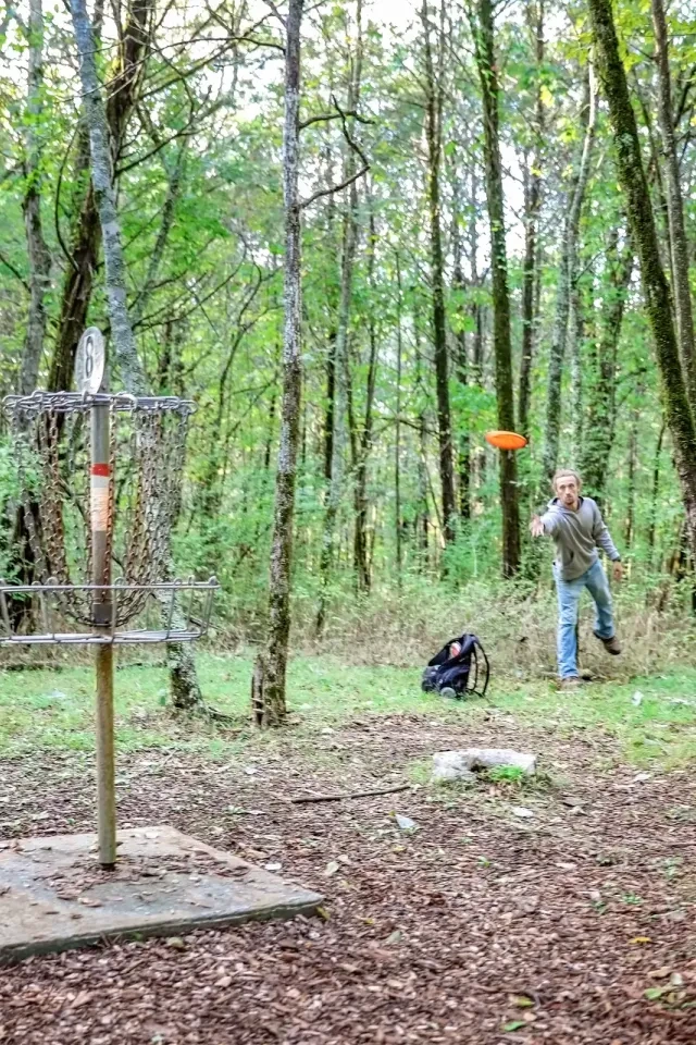 Person playing disc golf in a forest, throwing an orange disc toward a metal basket.