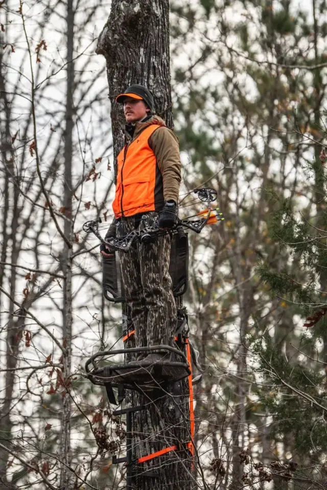 Hunter in orange vest standing on a tree stand, holding a bow in a forest.