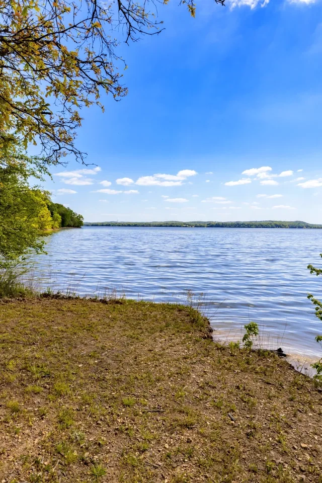 Lakeside view with trees, grassy shore, and blue sky with clouds.