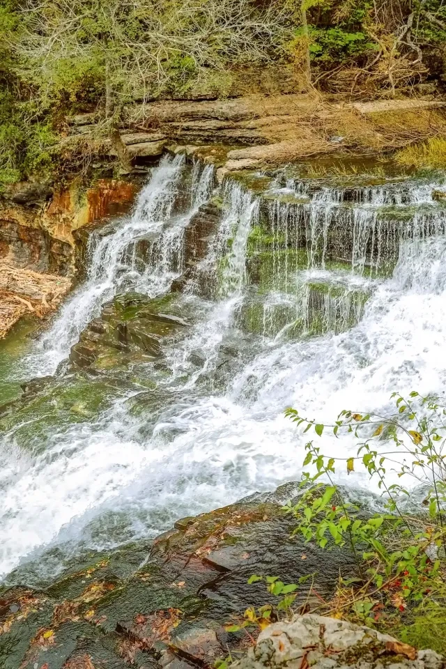 Waterfall cascading over rocks, surrounded by greenery and trees.