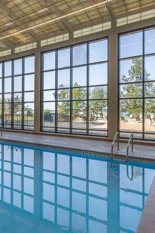 Indoor pool with large windows and outdoor view. Poolside chairs and tables nearby.