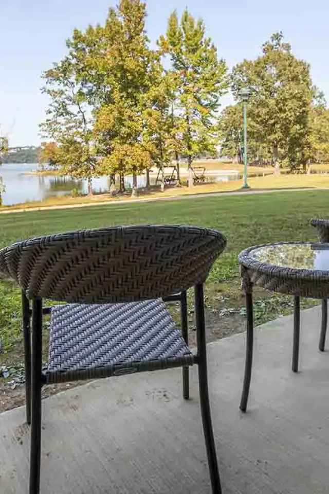 Two wicker chairs and a glass table on a patio overlooking a lake and trees.