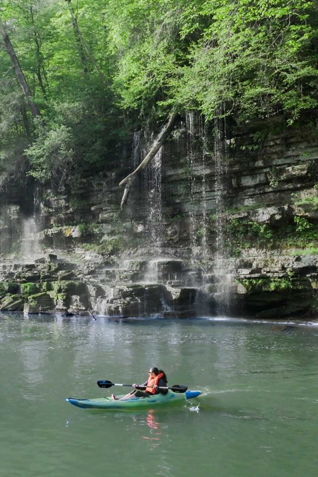 A person kayaking on a calm river near a mossy, rocky cliff with cascading waterfalls. Surrounding lush green foliage creates a serene atmosphere.