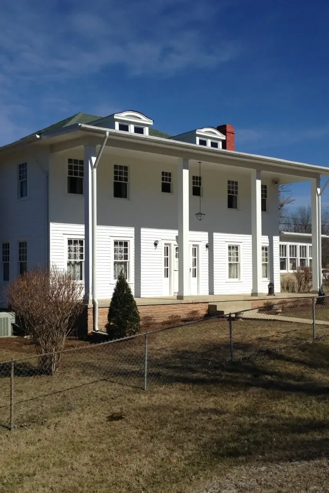 A large white colonial-style house with red brick chimneys stands under a bright blue sky, surrounded by a fenced grassy yard and bare trees.