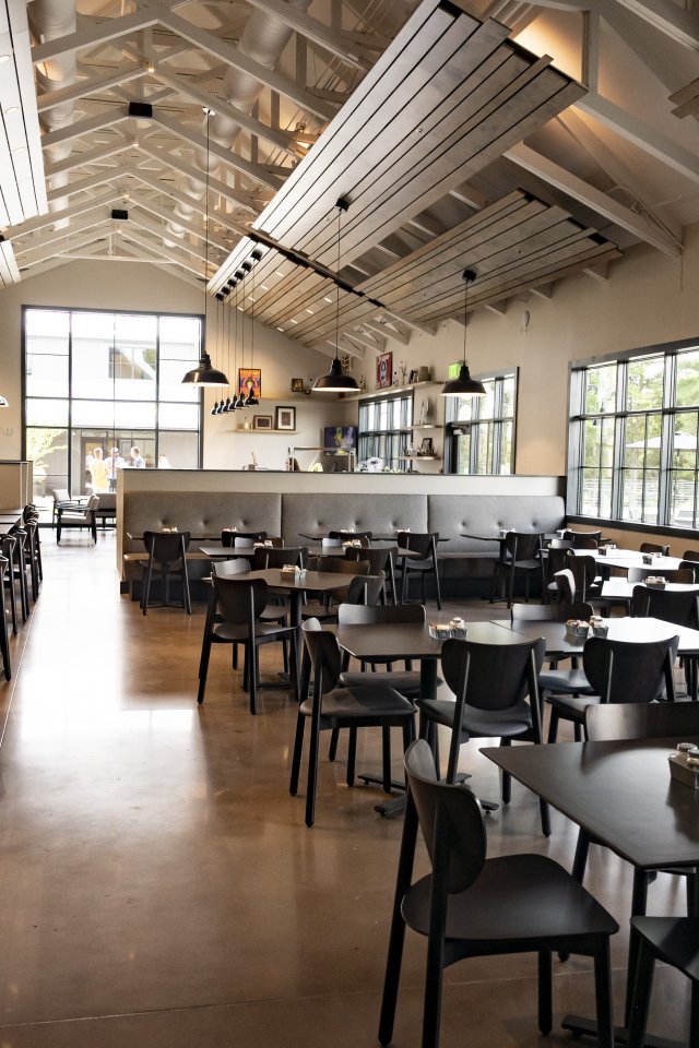 Spacious restaurant interior with high ceilings, modern black tables, and chairs. Large windows let in natural light, creating a calm, inviting atmosphere.