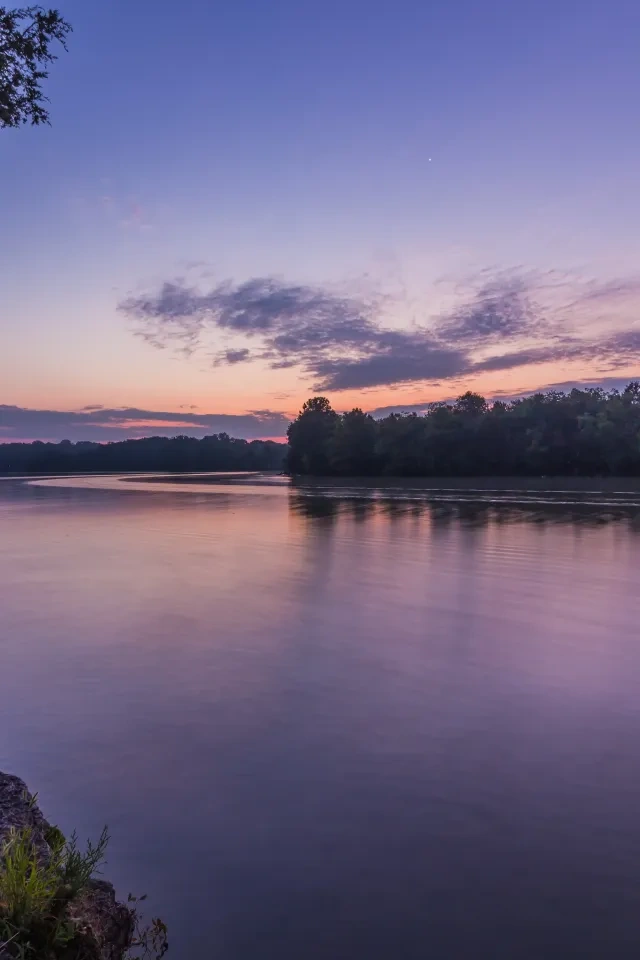 Calm river at sunset with trees and a purple sky.