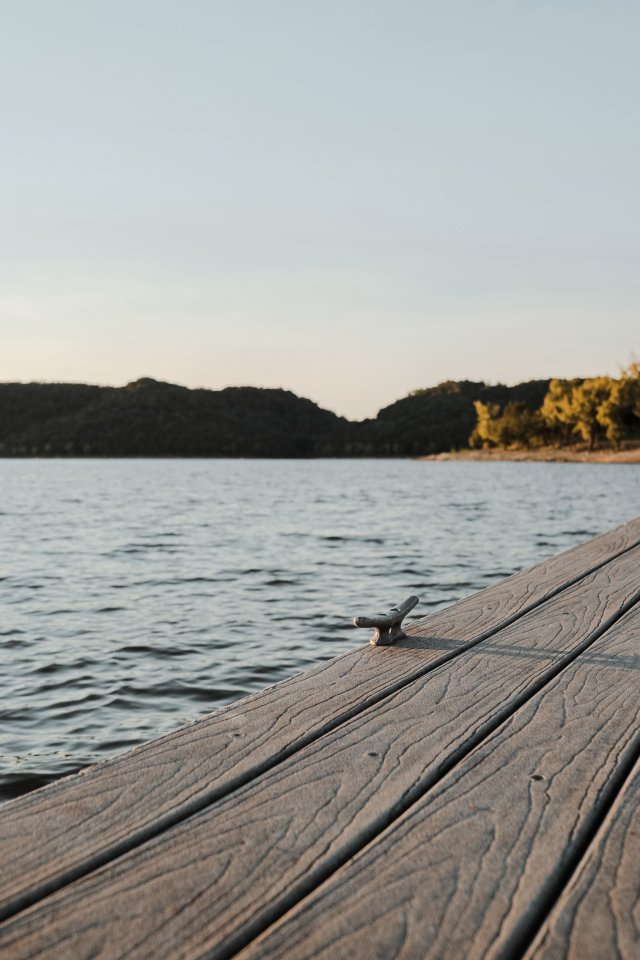 Wooden dock extending into a calm lake under a clear sky, surrounded by trees on distant hills, creating a peaceful, serene atmosphere at sunset.