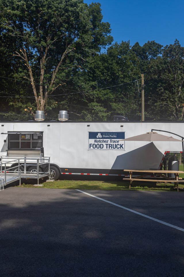 A white food truck in a park, shaded by tall trees and a clear blue sky. Nearby are picnic tables with umbrellas, creating a relaxed outdoor eating area.