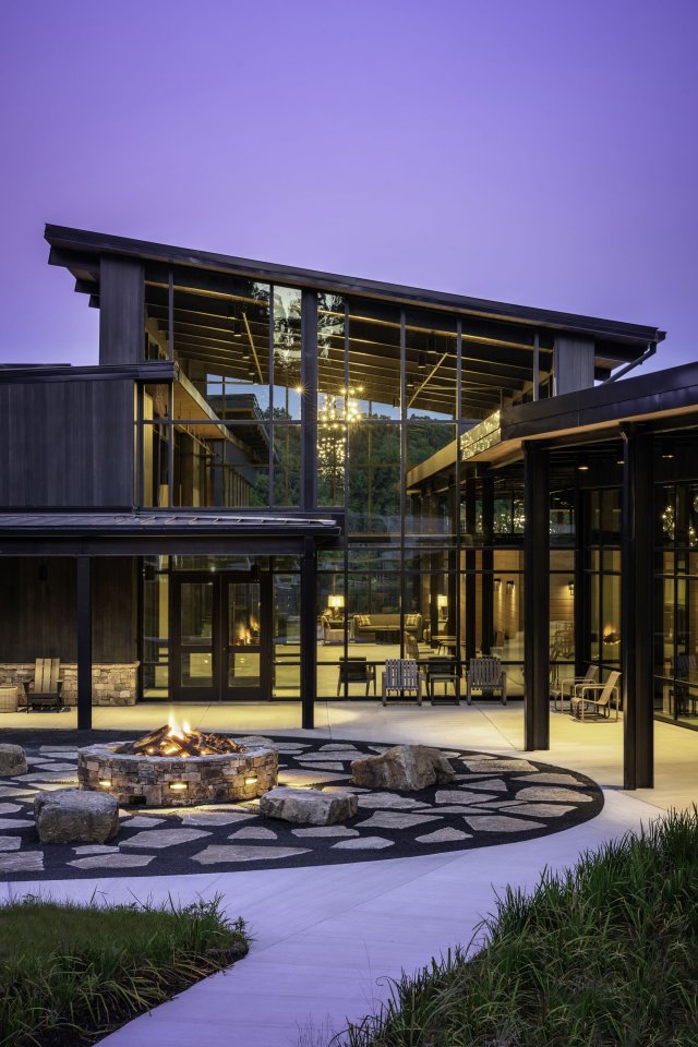 Modern building exterior at dusk with warm lights glowing through large glass windows. A circular fire pit is surrounded by stone seating.