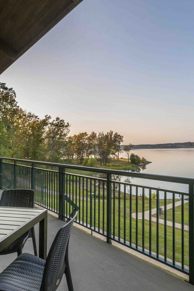 Covered patio with dark wicker tables overlooks a calm lake at sunset. Trees and a pier are visible, offering a peaceful, scenic view.