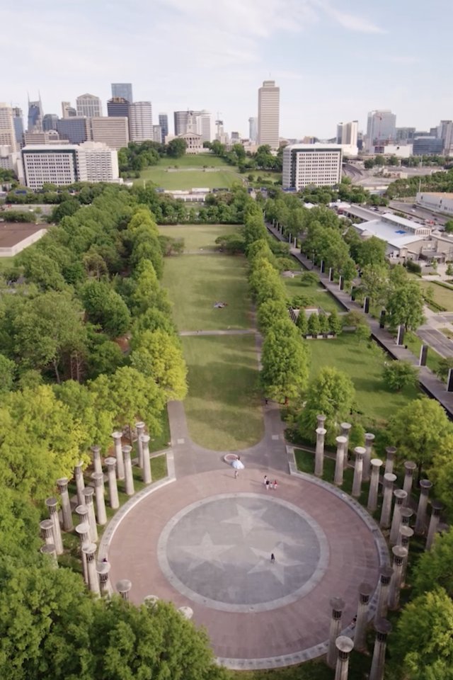 Aerial view of a city park with a circular plaza featuring a star pattern. Tree-lined paths extend towards a city skyline under a clear blue sky.