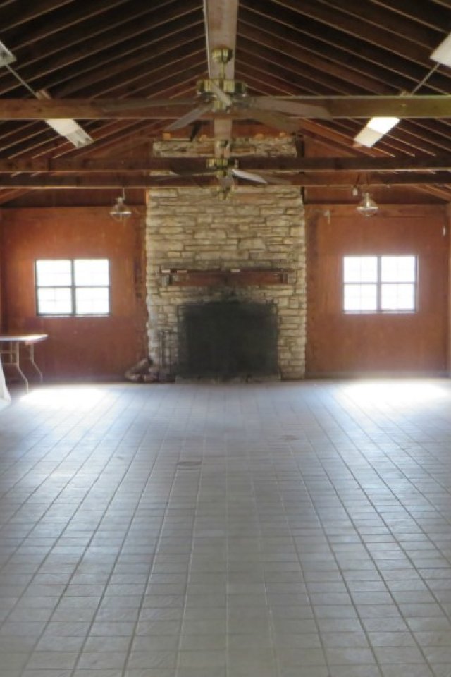 Rustic, empty wooden hall with tiled floors, stone fireplace at the end, exposed beam ceiling with fans, and windows along the sides, creating a serene, cozy atmosphere.