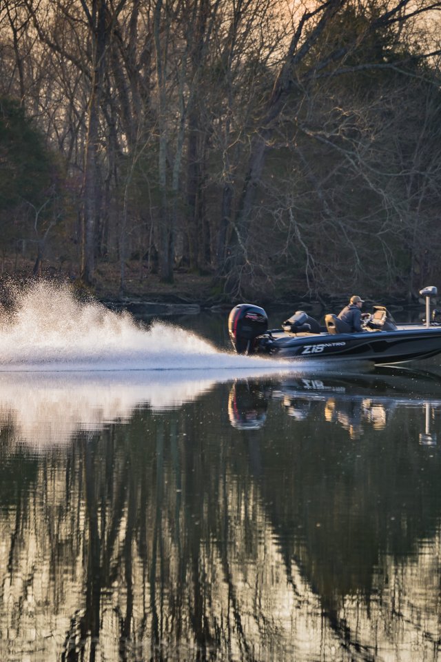 A speedboat creates a trail of white spray as it skims across a calm lake, surrounded by bare trees, reflecting a serene winter scene at dusk.