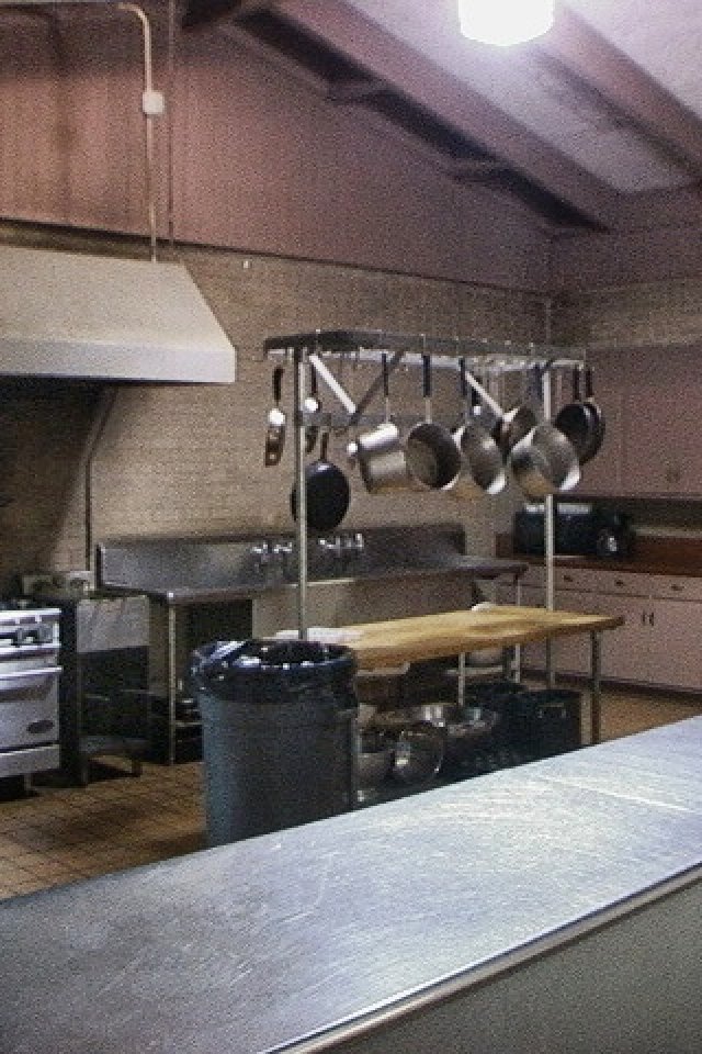 A spacious industrial kitchen with a metal stove, overhead exhaust, and hanging pots. A large prep table and stainless steel counters create a functional, clean atmosphere.