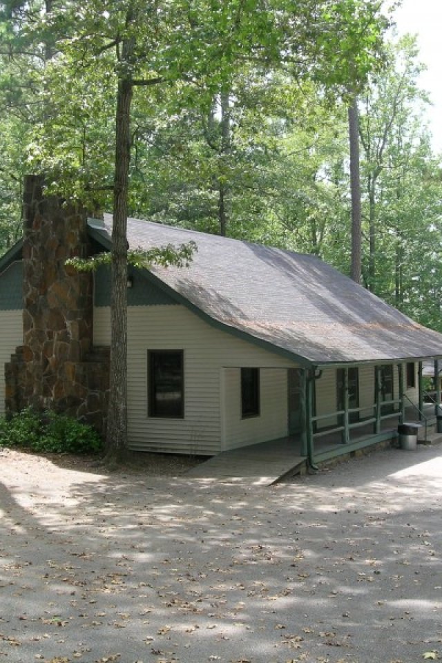 A rustic cabin with a stone chimney nestled among tall trees. The setting is serene, with sunlight dappling the roof and leafy shadows on the ground.