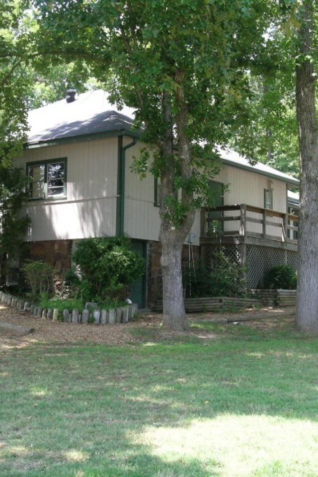 A cozy, single-story house with a light gray exterior and a sloped roof is nestled among tall trees, with a wooden ramp leading to the entrance. Sunlight filters through the leaves, casting dappled shadows on the grassy yard, creating a serene, inviting atmosphere.
