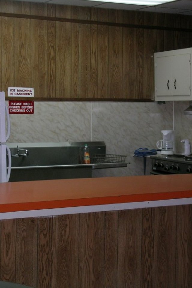 A retro kitchen with wood-paneled walls shows an orange countertop, white fridge, and microwave. A stove and sink with a soap dispenser are visible.
