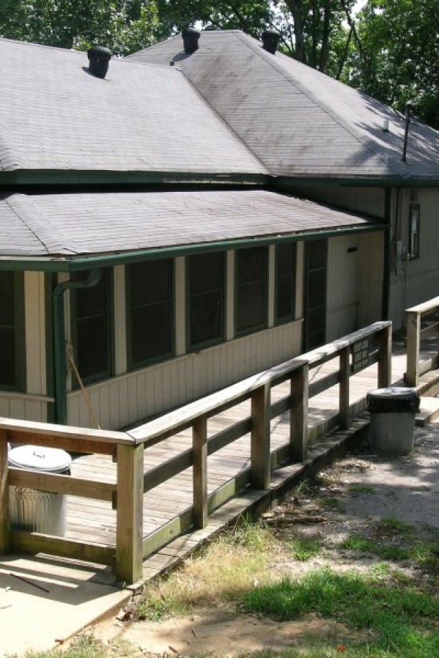 A cozy, single-story house with a light gray exterior and a sloped roof is nestled among tall trees, with a wooden ramp leading to the entrance. Sunlight filters through the leaves, casting dappled shadows on the grassy yard, creating a serene, inviting atmosphere.