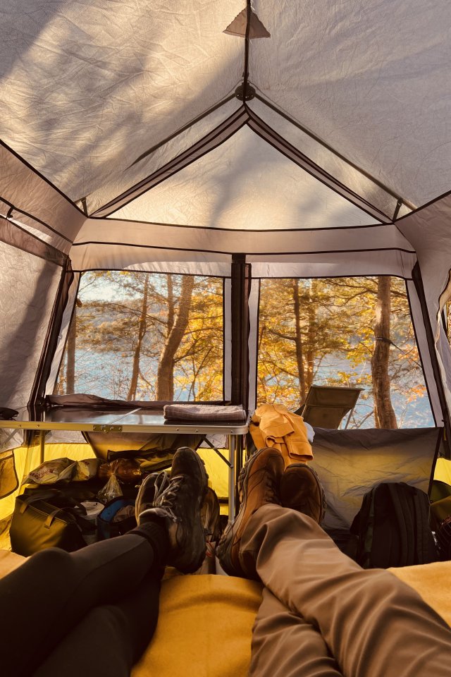 A person relaxes in a hammock near a campfire, surrounded by autumn trees. A lake reflects the warm sunset, creating a serene and peaceful atmosphere.