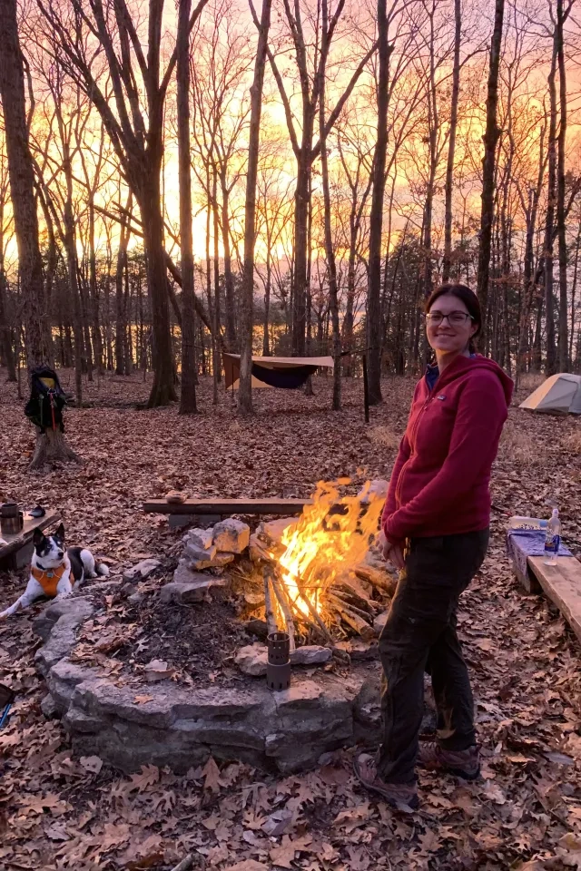 Smiling person by a campfire in a wooded area at sunset.