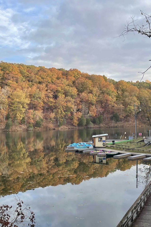 Autumn scene with a calm lake reflecting vibrant orange and green trees. A lone person walks on a wooden boardwalk by the water.