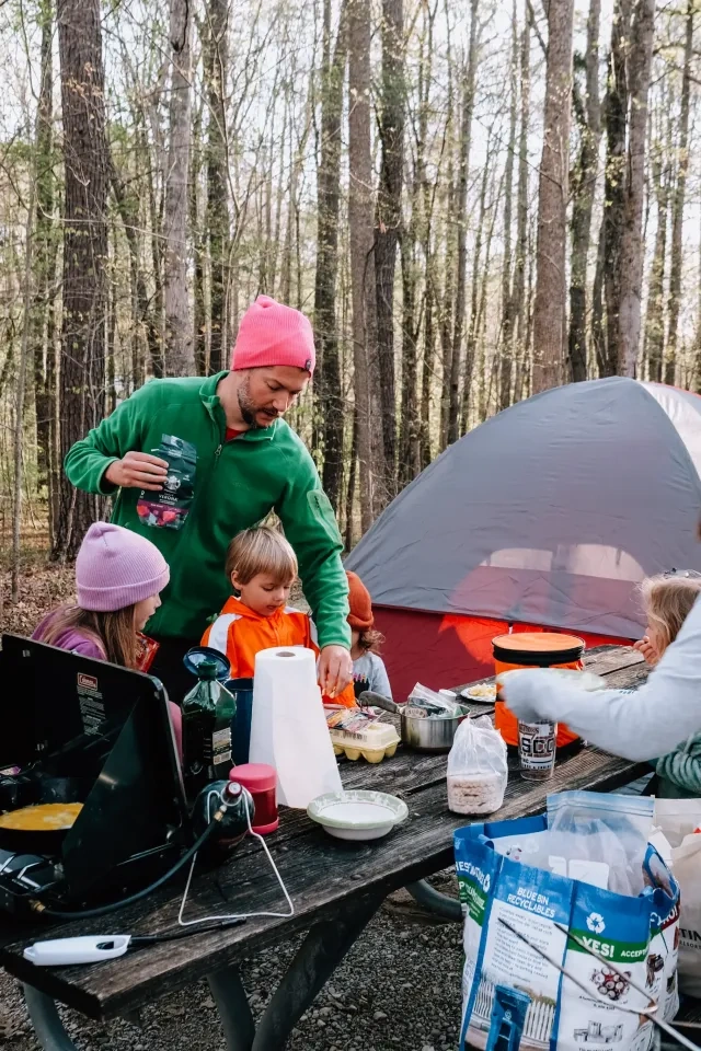 Family cooking at a campsite with a tent and trees in the background.