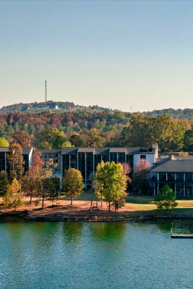 Lakeside resort buildings surrounded by lush trees with a hill in the background.