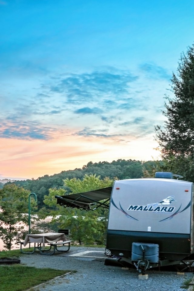Travel trailer parked by a lake at sunset, surrounded by trees.