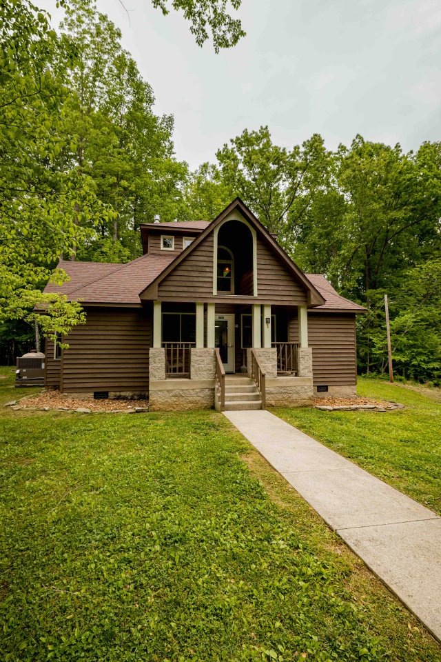 Brown wooden cabin with a gabled entrance and a pathway in a forest setting.