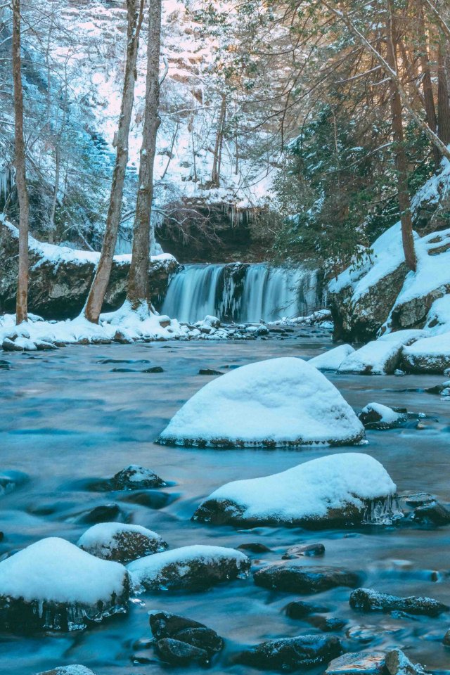 Snow-covered river with rocks and a small waterfall in a forest.