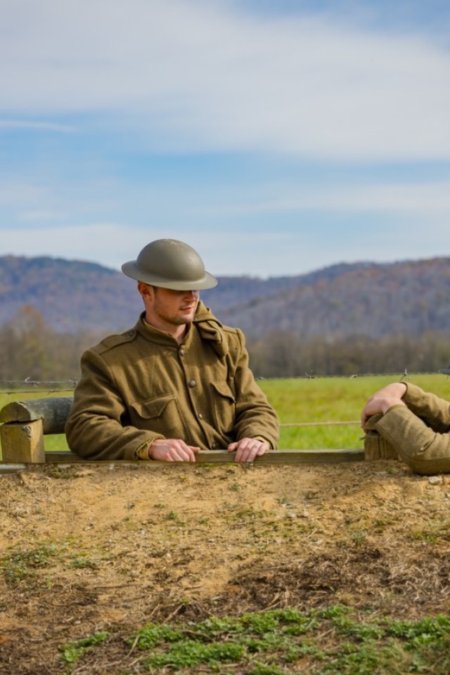 Two men dressed as World War One soldiers stand face to face in a trench in a bright green field with a hazy view of mountains behind them. 
