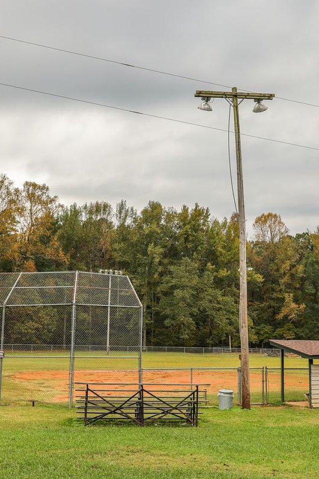 Baseball field with bleachers, dugout, and trees in the background. Overcast sky.