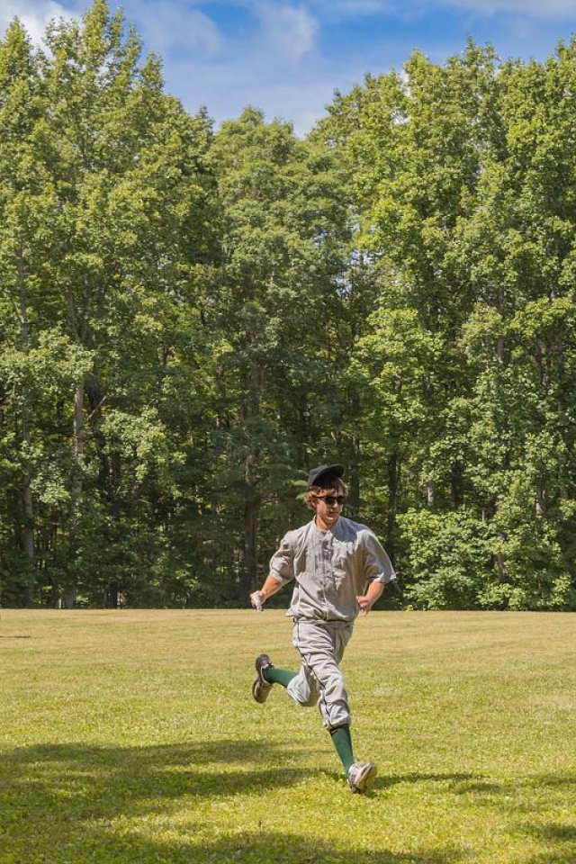 Baseball players running on a grassy field with trees in the background.