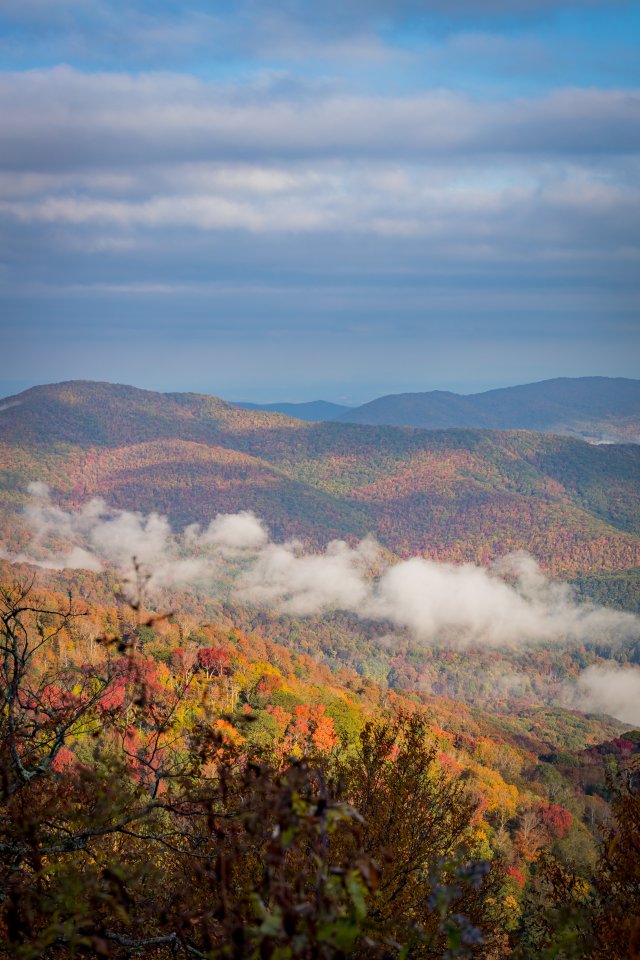 Scenic view of layered mountain ridges covered in vibrant fall foliage, with trees displaying shades of red, orange, yellow, and green. Low clouds or fog drift through the valleys between the hills, while a partly cloudy blue sky stretches above.