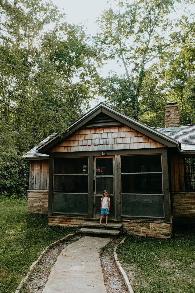 Child standing in front of a rustic cabin surrounded by trees.