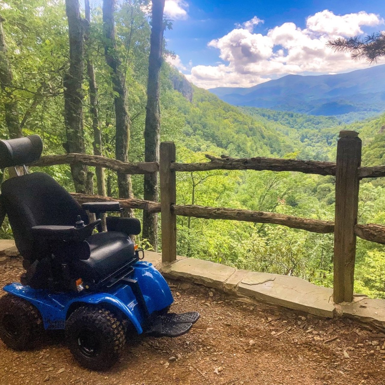 A blue all-terrain wheelchair is parked on a dirt path overlooking a lush green mountain valley.