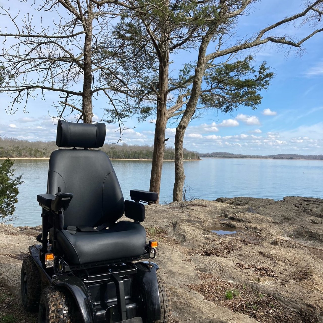 A black mobility scooter is parked on a rocky lakeside, surrounded by trees.