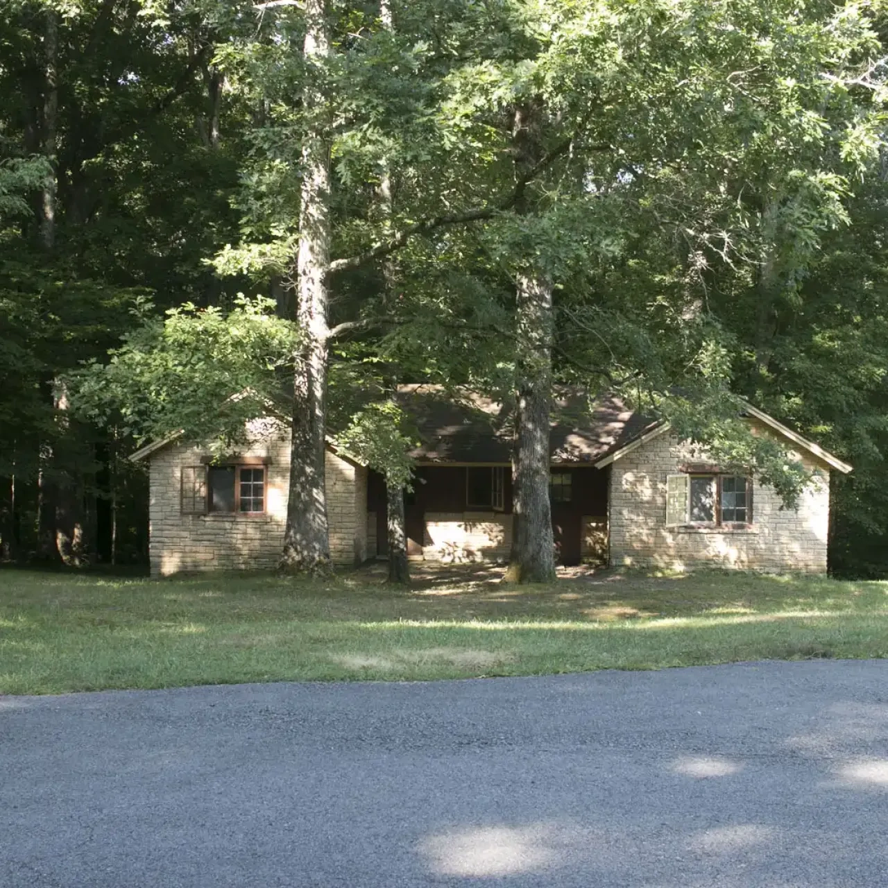 Stone house in a forest, shaded by large trees, with a grassy yard in front.